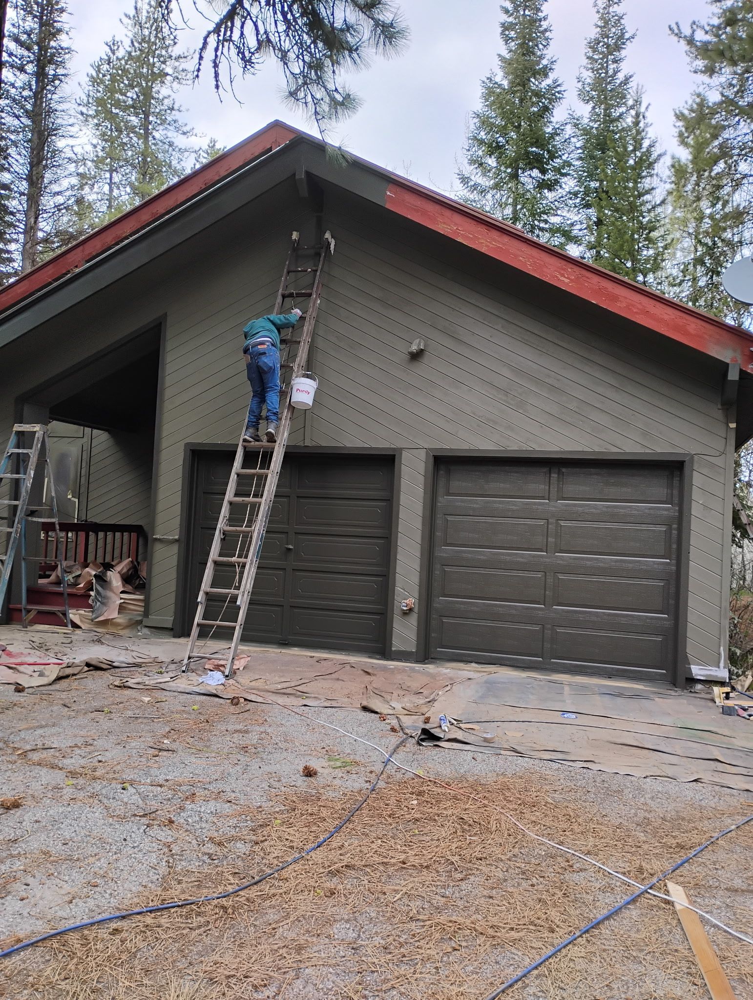 Worker on a ladder painting a house gable above two garage doors in a wooded driveway