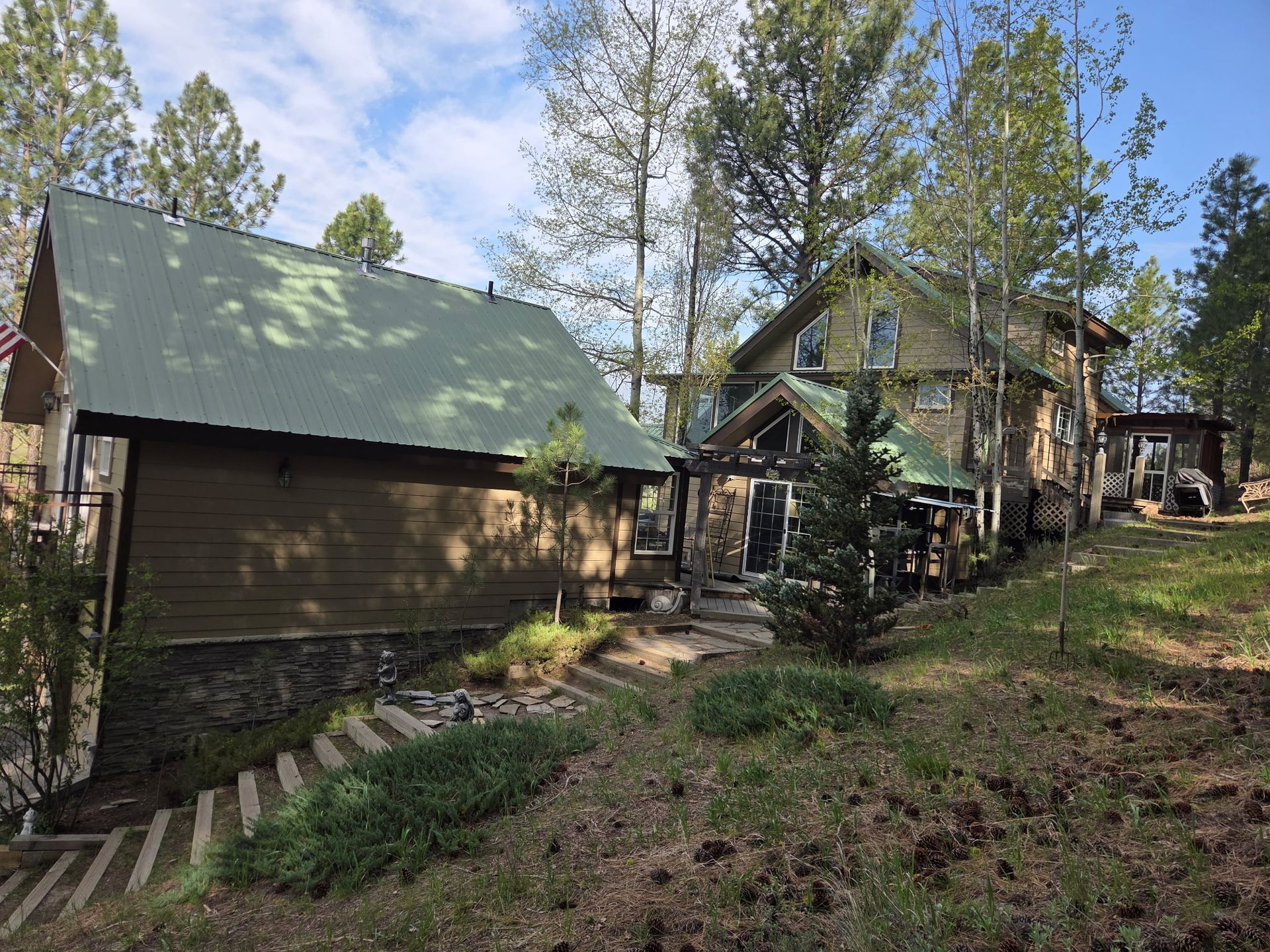 Wooded cabin with green roof and stone chimney on a sloped hillside under blue sky