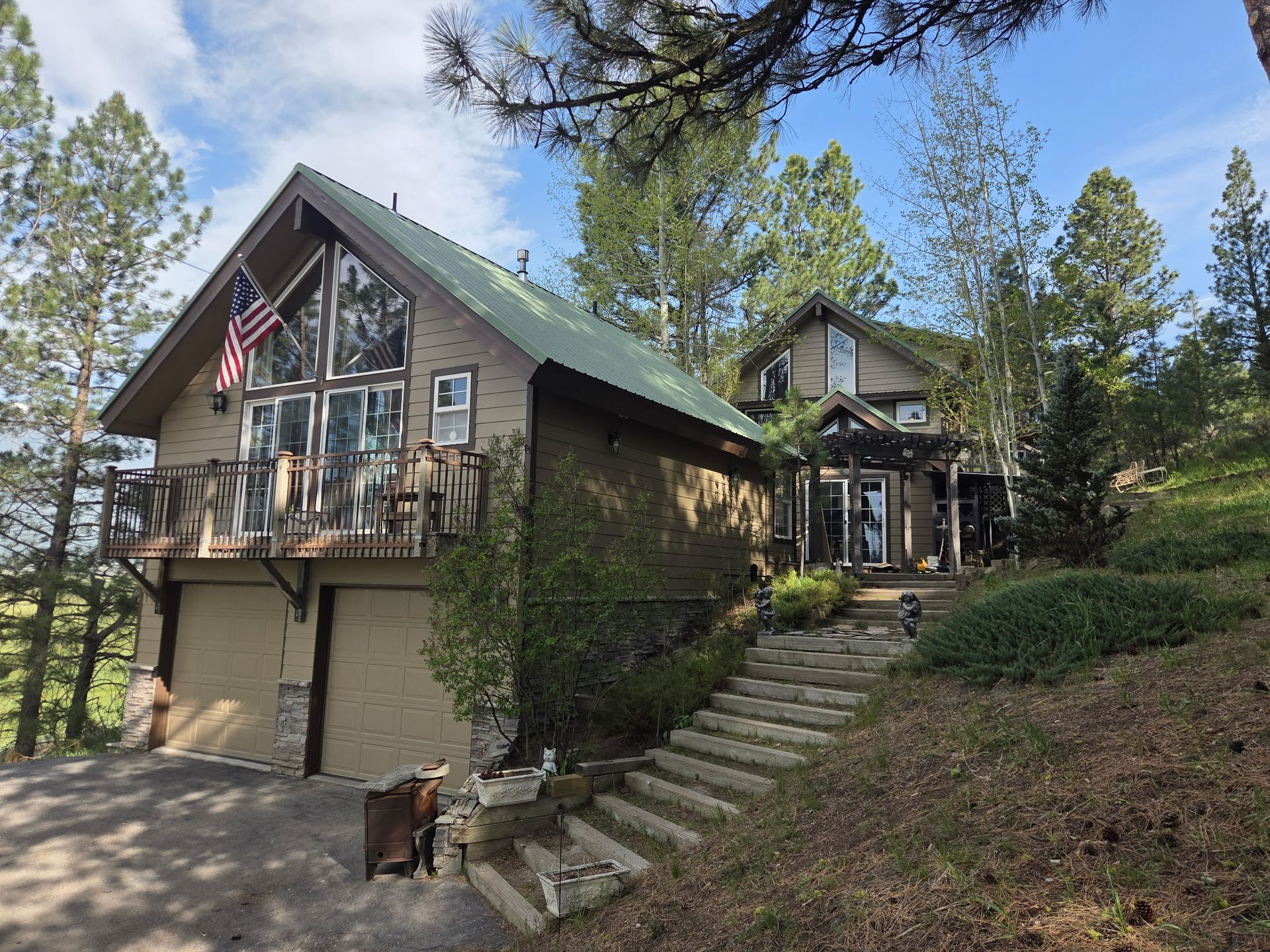 Cabin-style house with stone stairs, balconies, and trees on a wooded hillside under blue sky