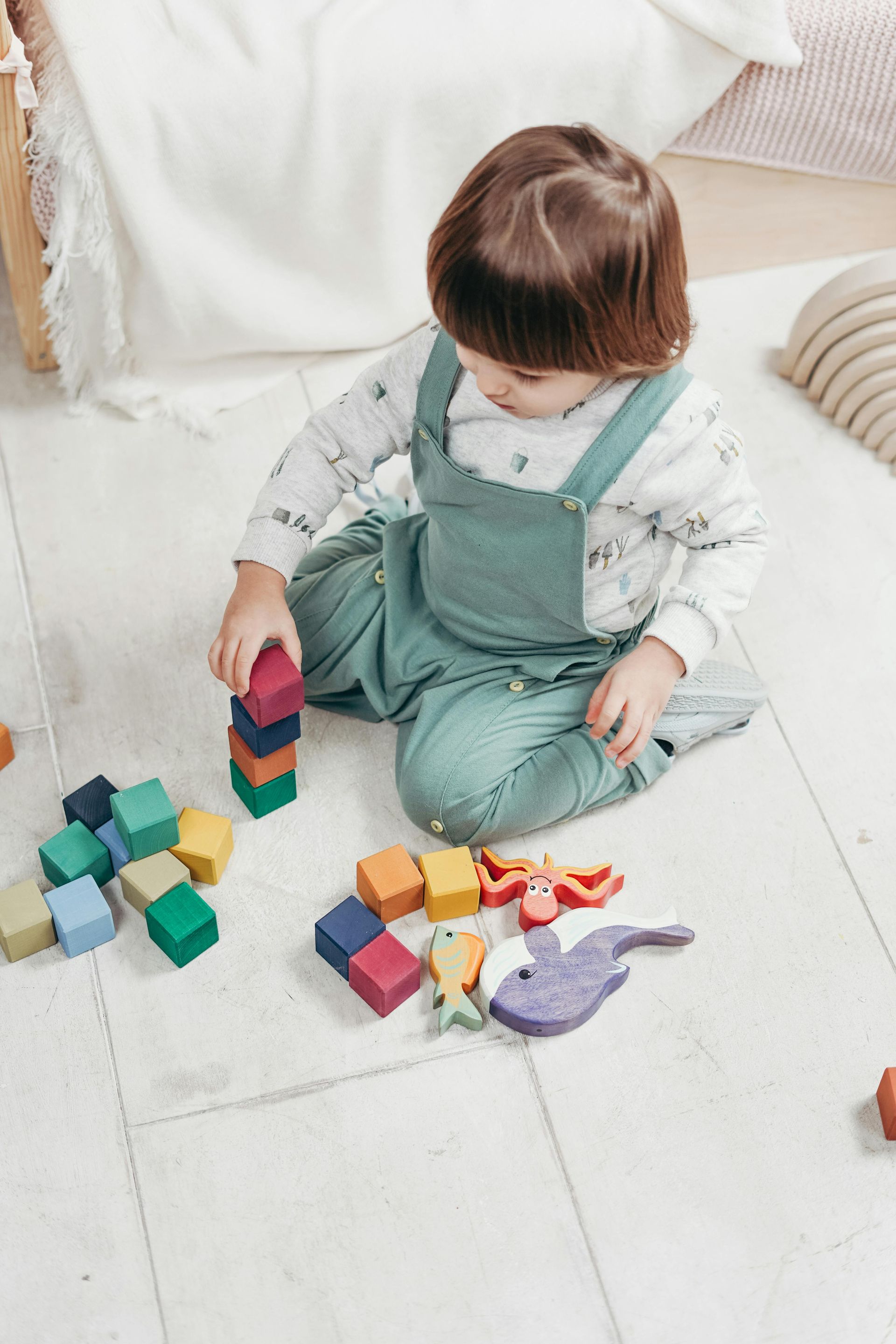 Child in green overalls playing with colorful wooden blocks on a white tiled floor.