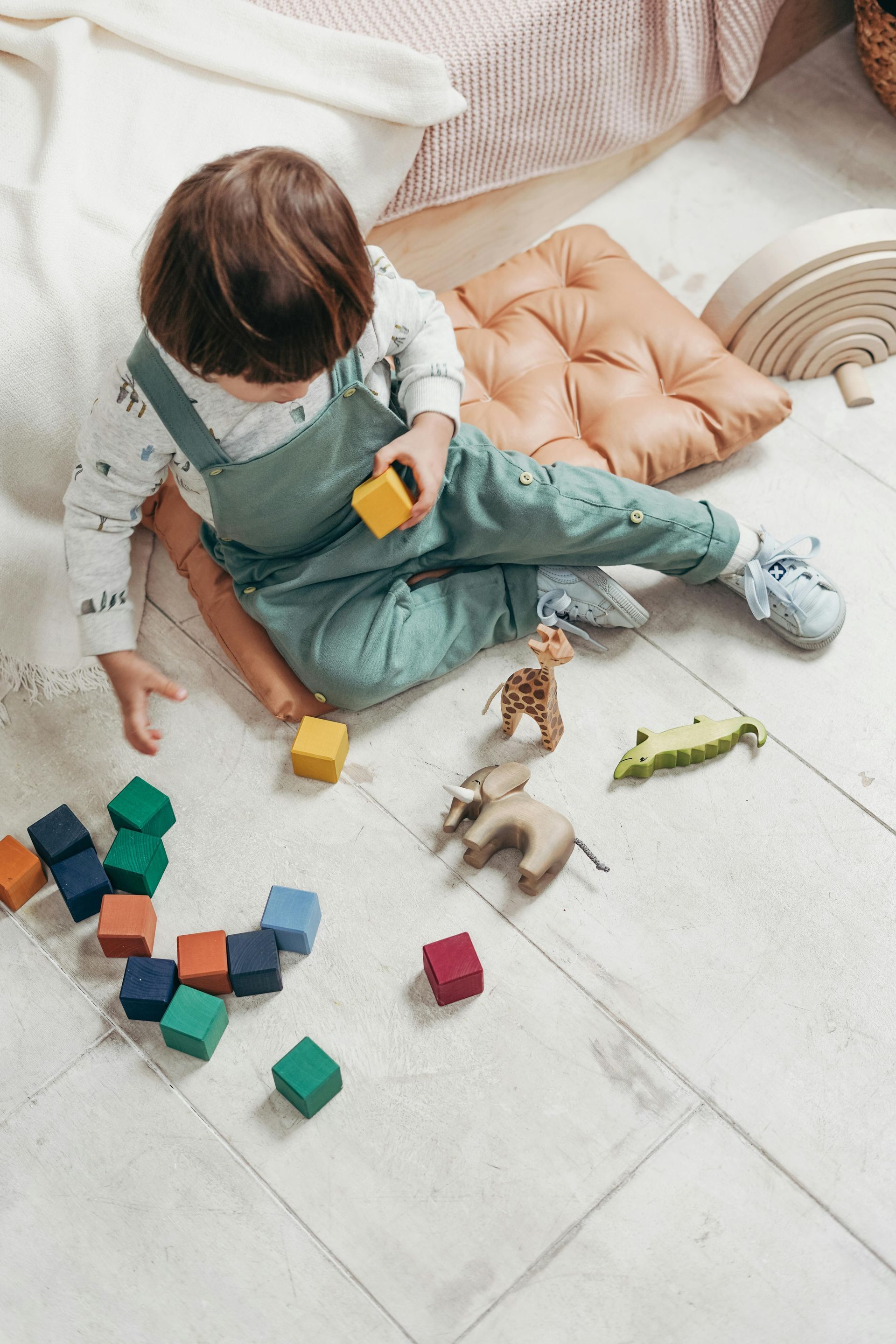 Child in overalls playing with blocks and animal toys on a light floor.
