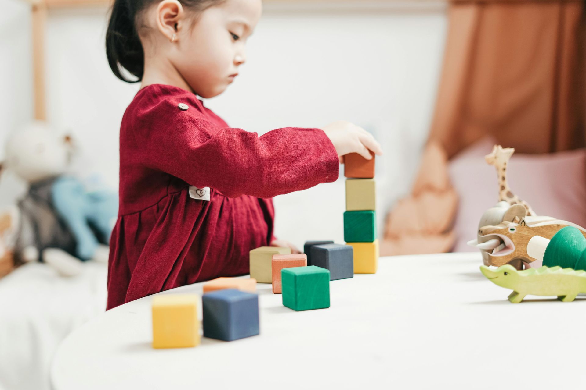 Young girl in red dress stacking colorful blocks on a white table.