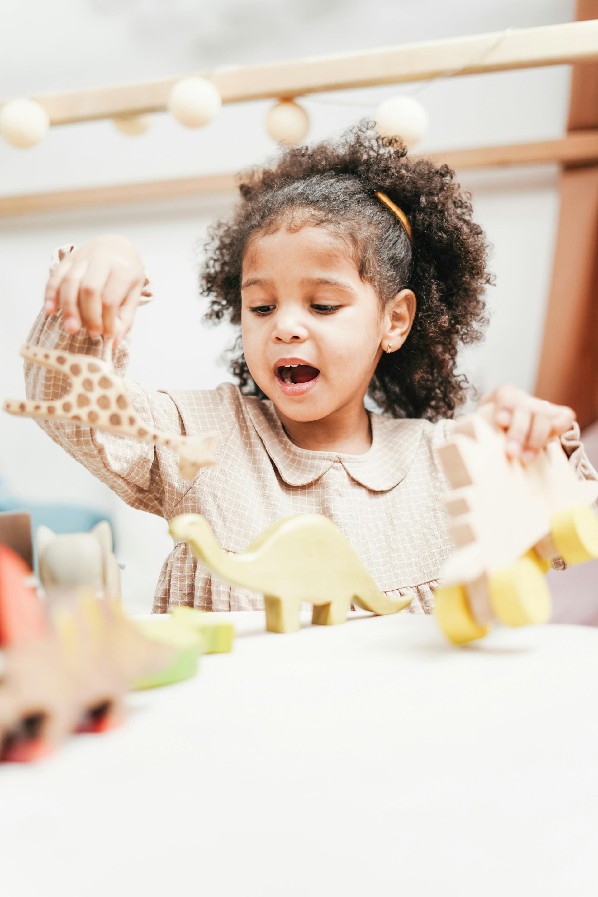 Young girl with curly hair plays with wooden dinosaur toys at a table.