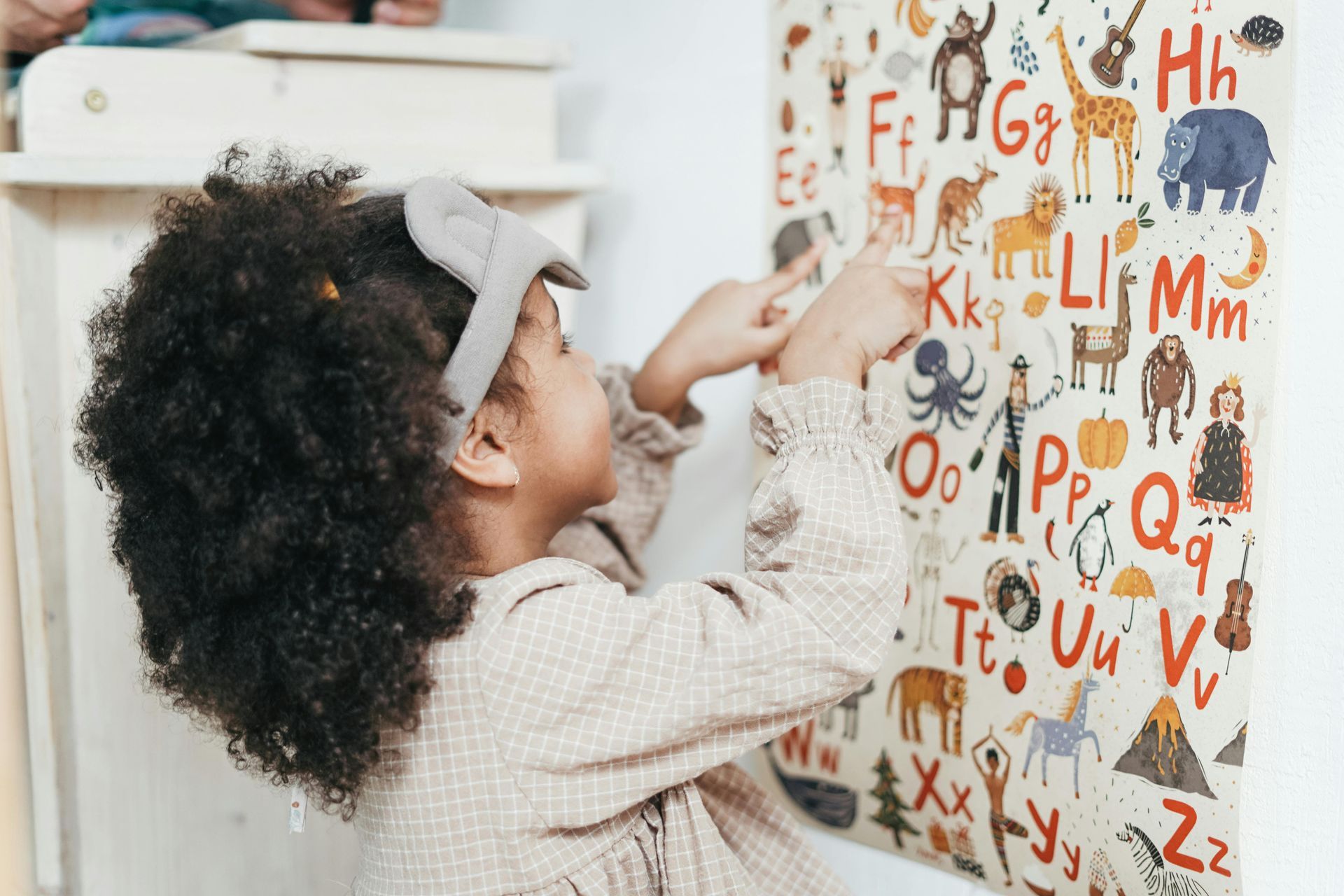 Girl with curly hair pointing at an alphabet poster on a white wall.