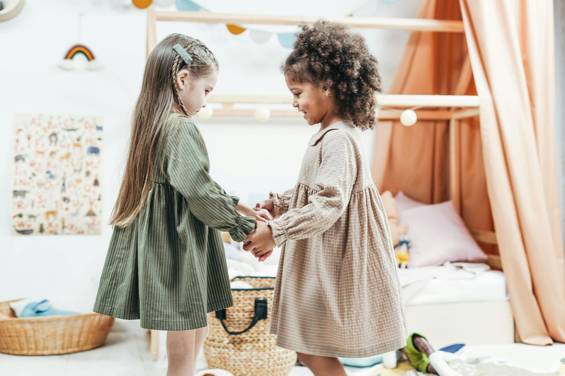 Two young girls holding hands, smiling, in a nursery room with a bed and toys.