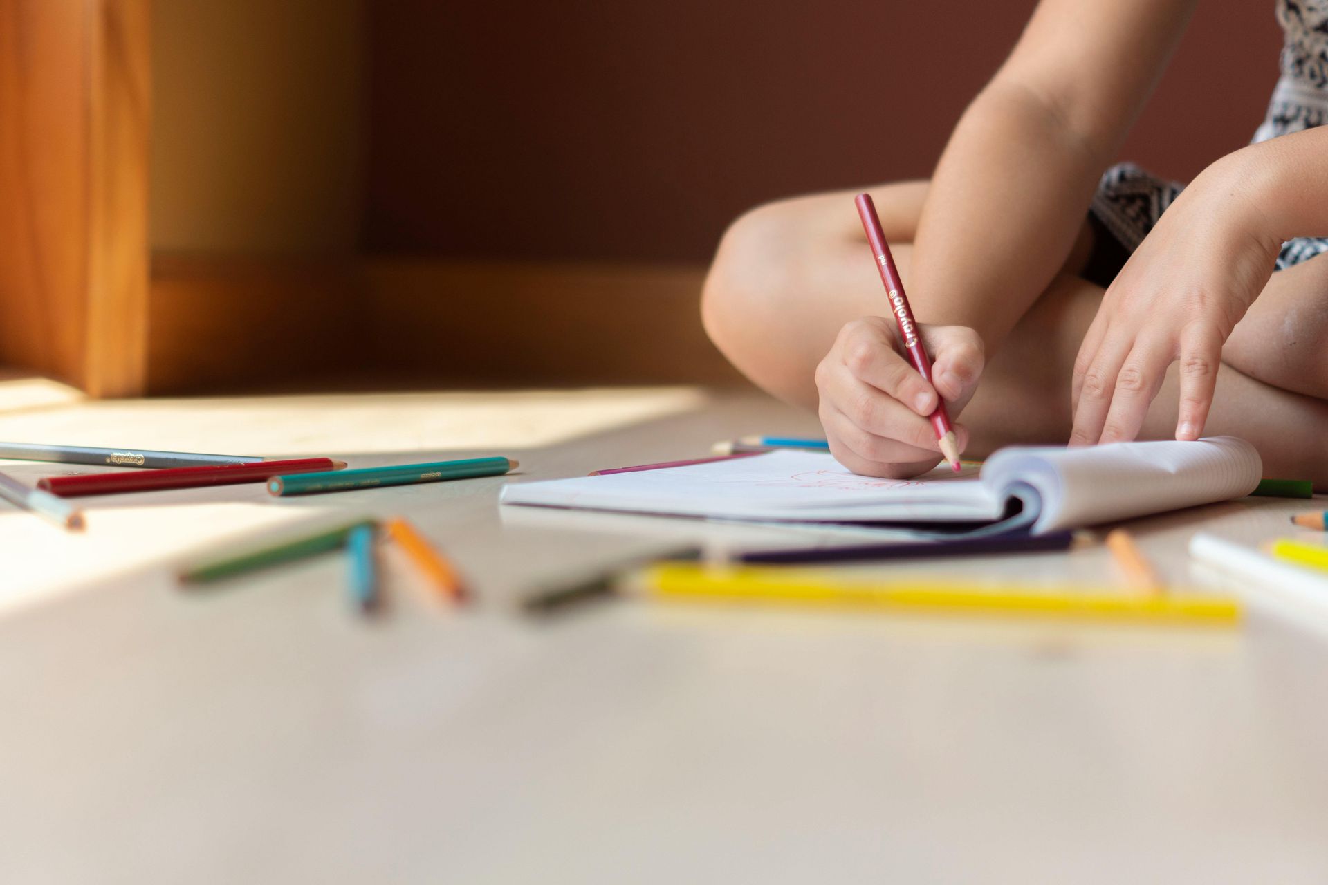 Child sitting on the floor drawing with a red pencil, surrounded by scattered colored pencils.