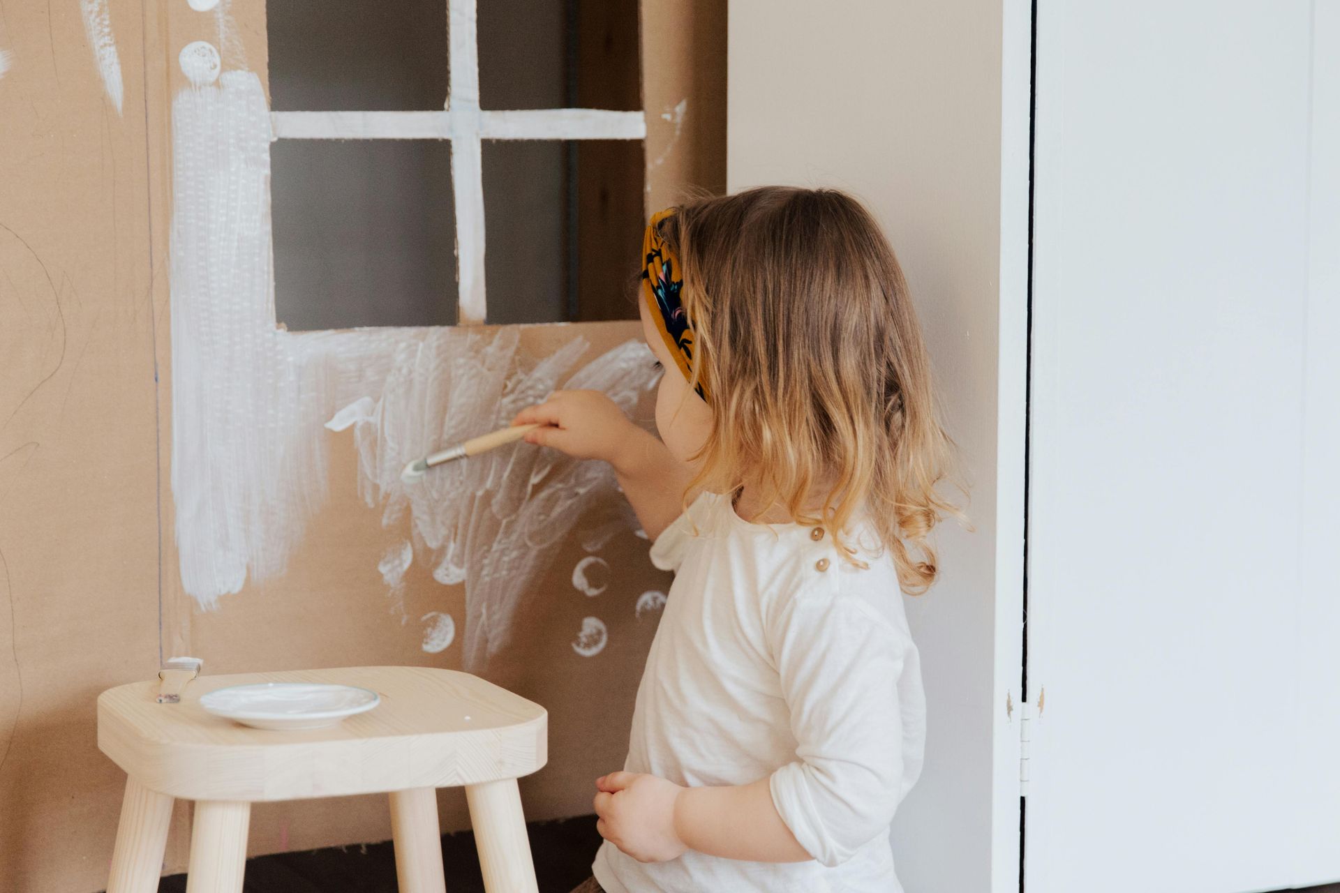 A child with messy hair paints a cardboard window with white paint.