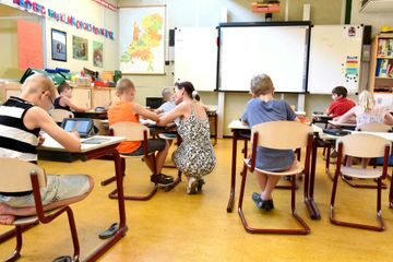 School classroom with students at desks, teacher assisting, map and whiteboard on the wall.