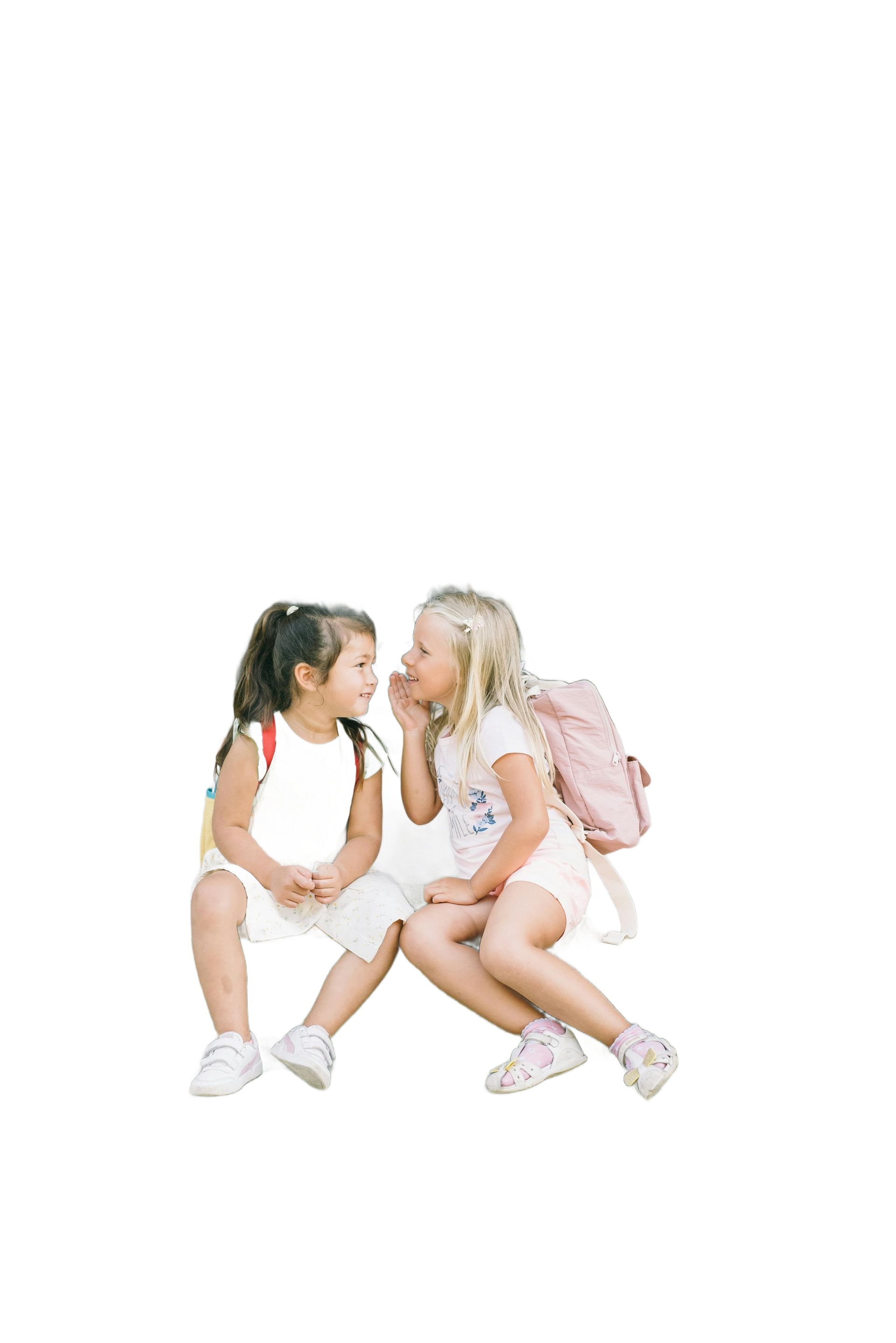 Two young girls whispering; one blonde with a pink backpack, the other dark-haired. Outdoors, white clothing.