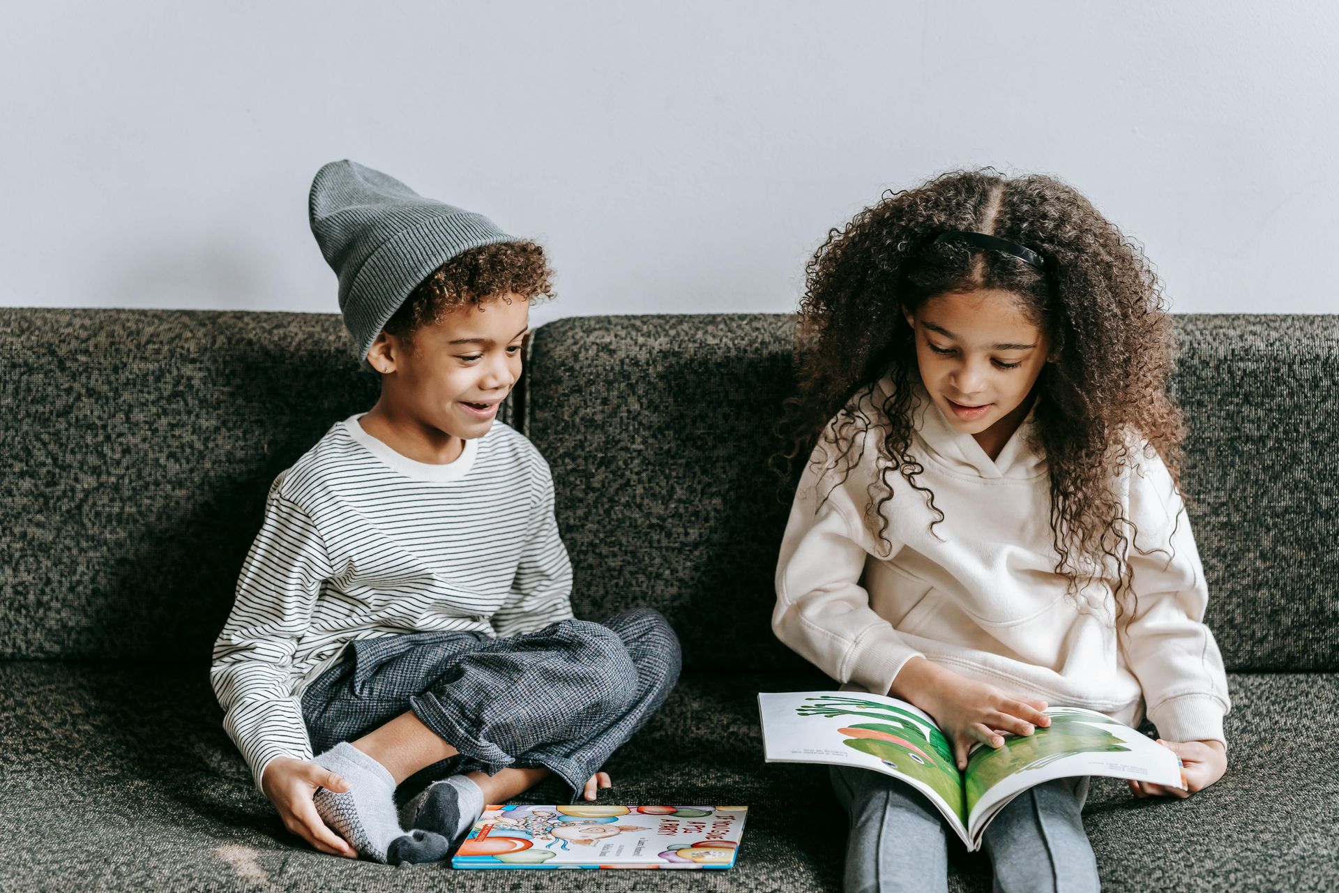 Two children on a couch reading books, one wearing a hat, smiling.