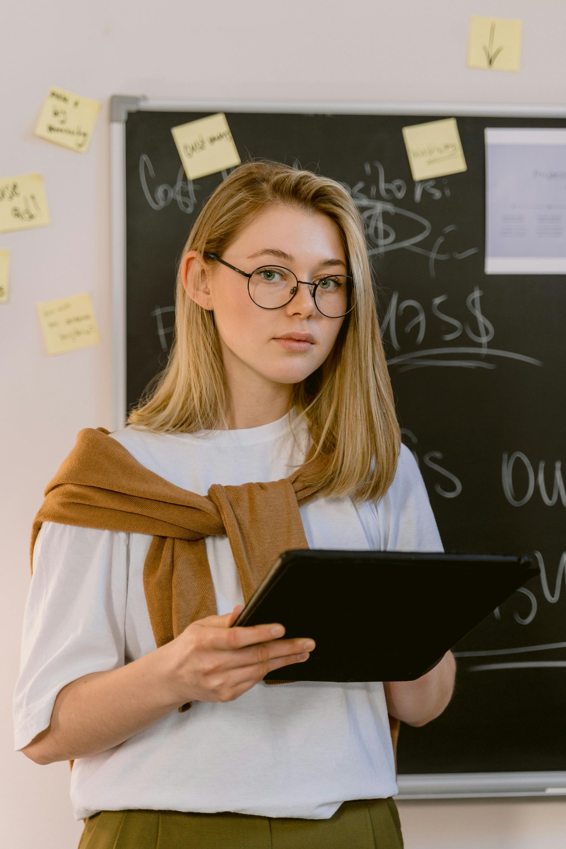 Woman with glasses, tablet, and sweater stands in front of a blackboard, possibly teaching or presenting.