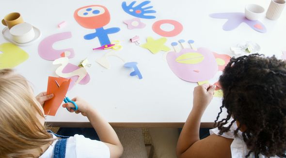 Two children at a table crafting colorful paper shapes with scissors.