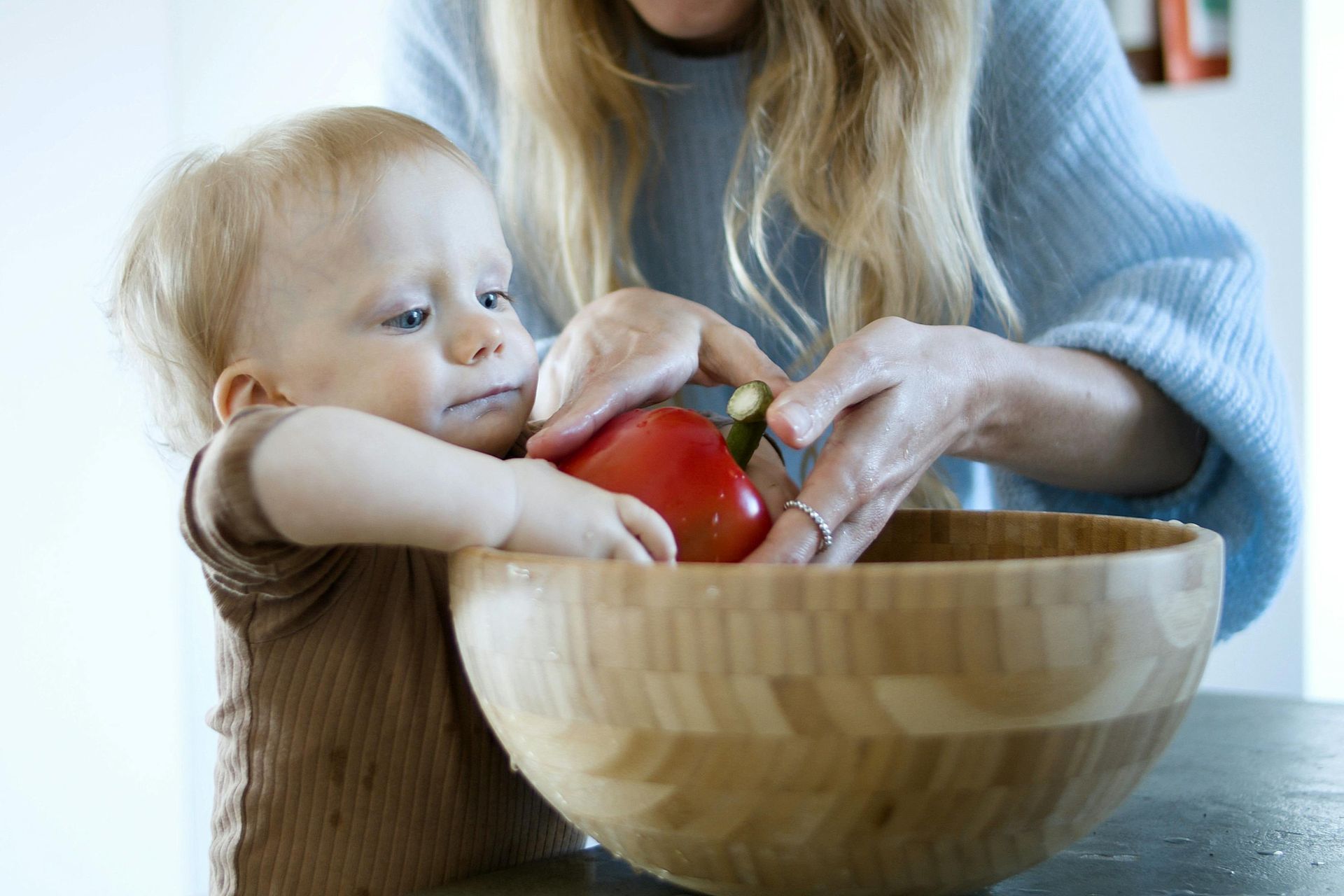 Baby and adult in kitchen with red pepper in wooden bowl.