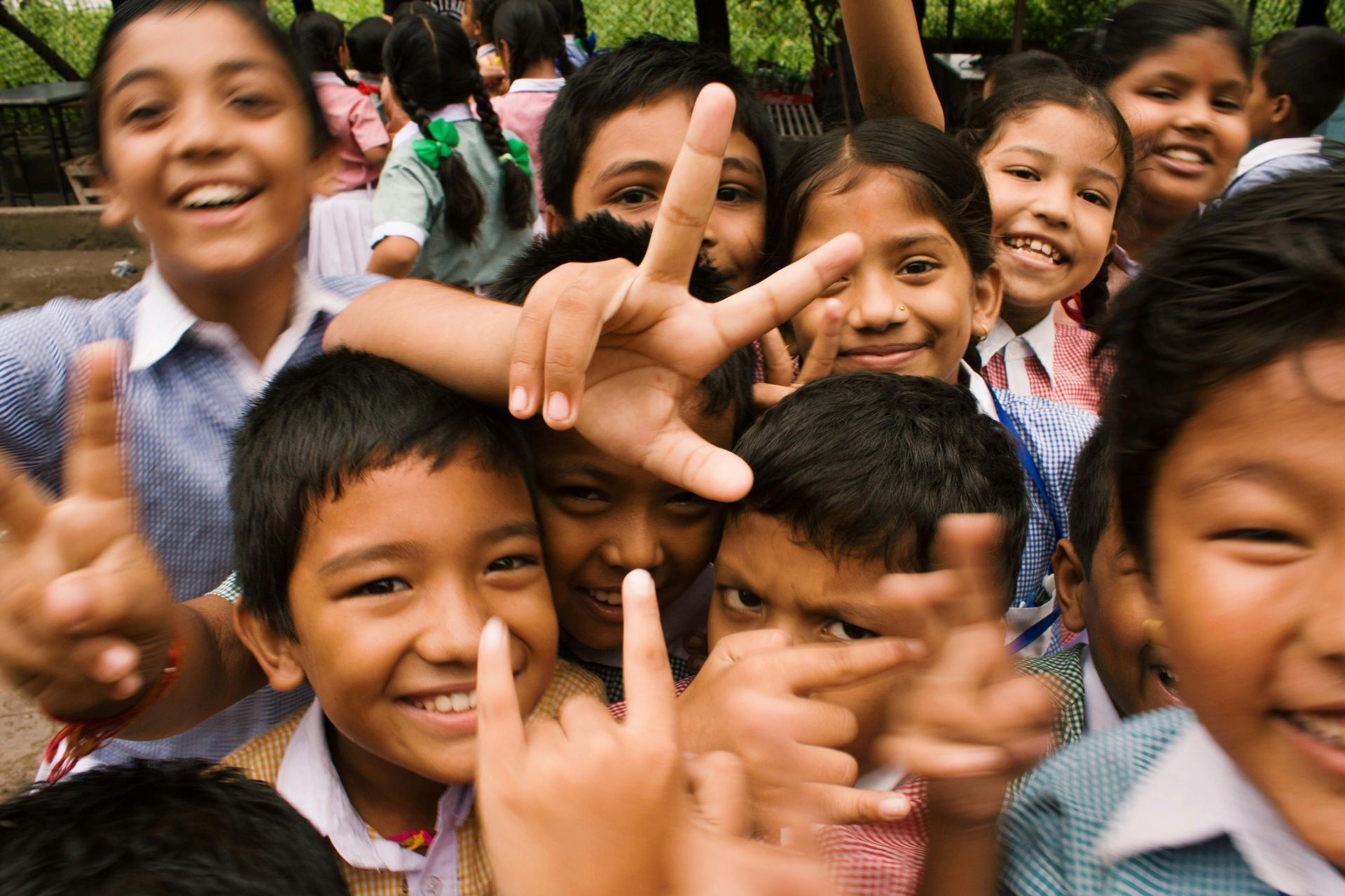 Group of smiling children, possibly school students, outside. They are making hand gestures and appear joyful.