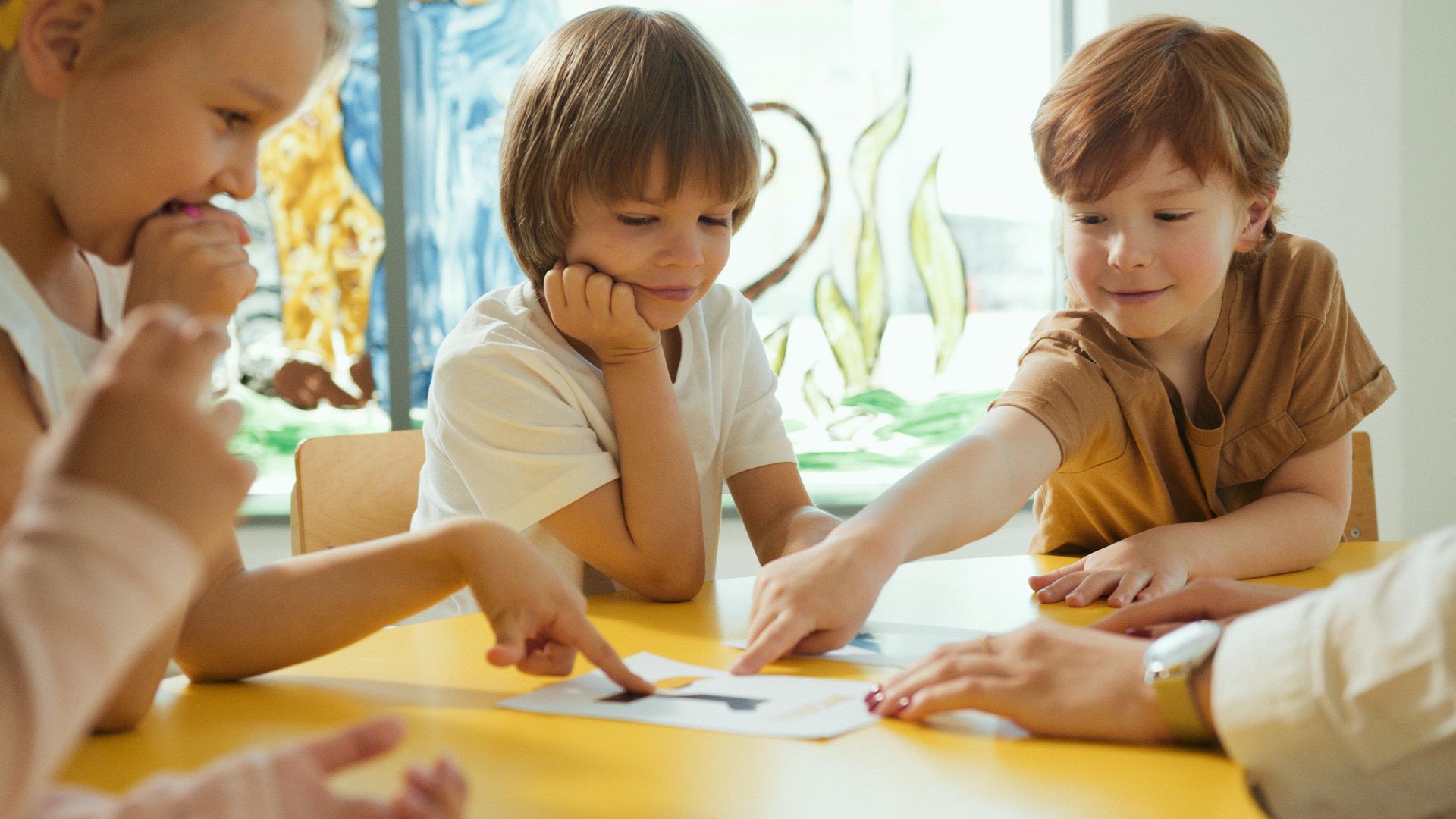 Children at a yellow table, looking at papers with an adult, in a bright room.