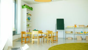 Brightly lit children's classroom with tables, chairs, shelves, and a green rug.