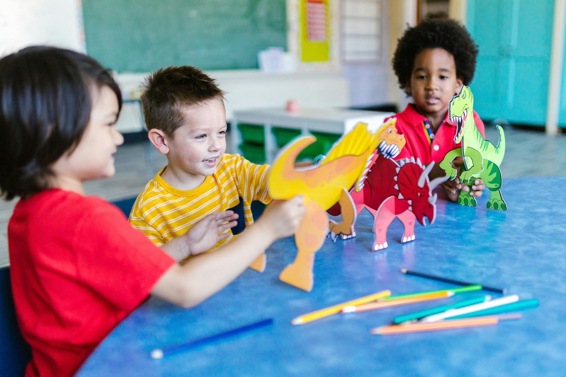 Three diverse children playing with colorful dinosaur cutouts at a blue table in a classroom.