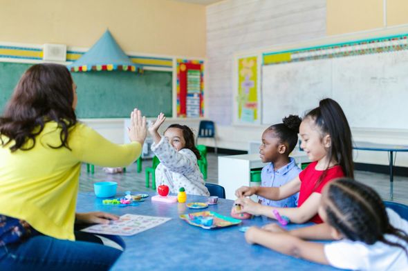 Teacher high-fives a student in a colorful classroom, surrounded by other children at a table.