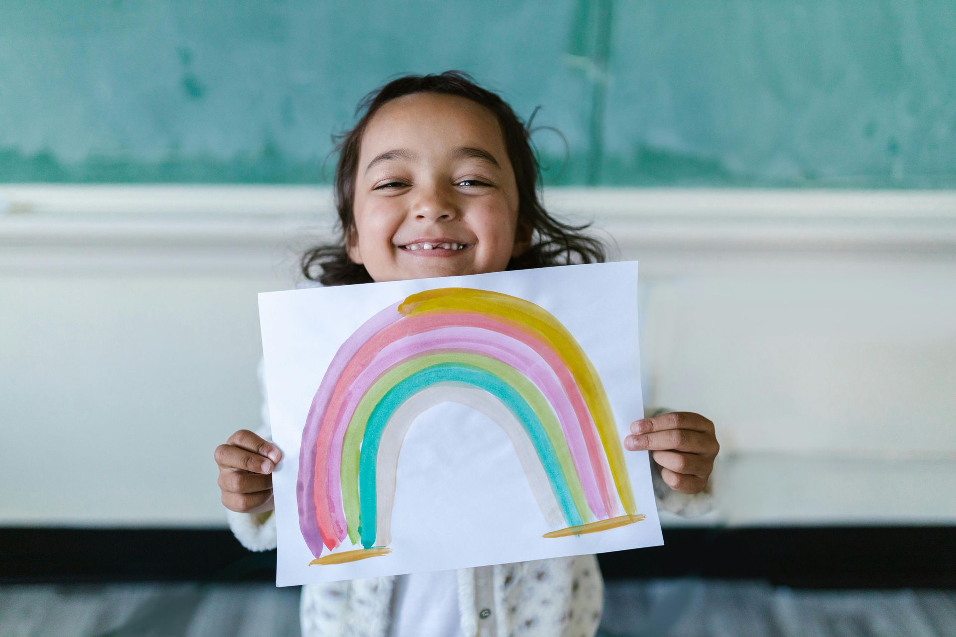 Girl smiles, holding up a watercolor rainbow drawing in front of a green chalkboard.