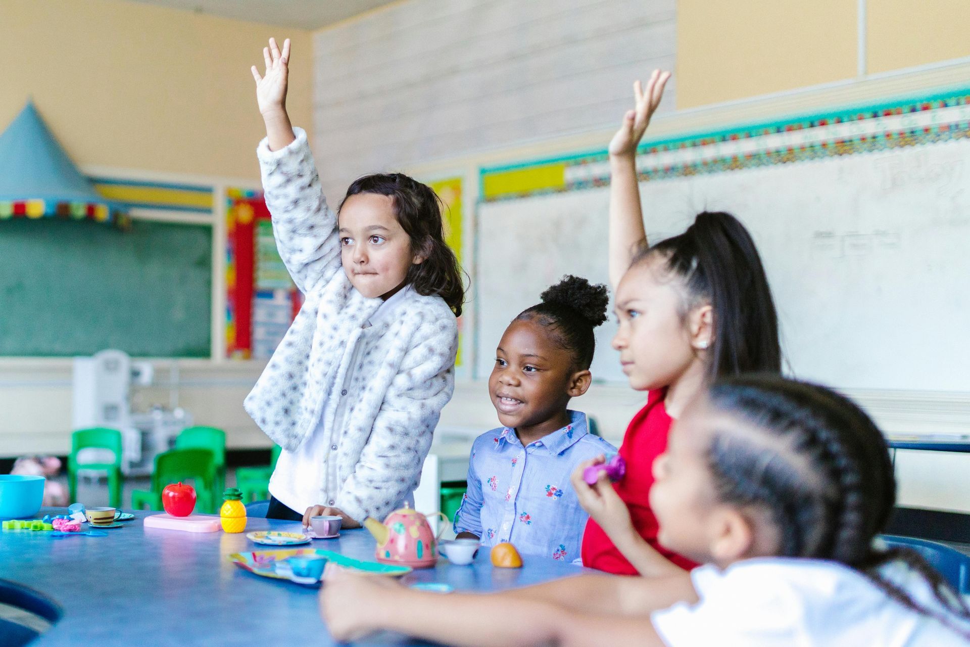 Children raising hands in a classroom, looking at something.