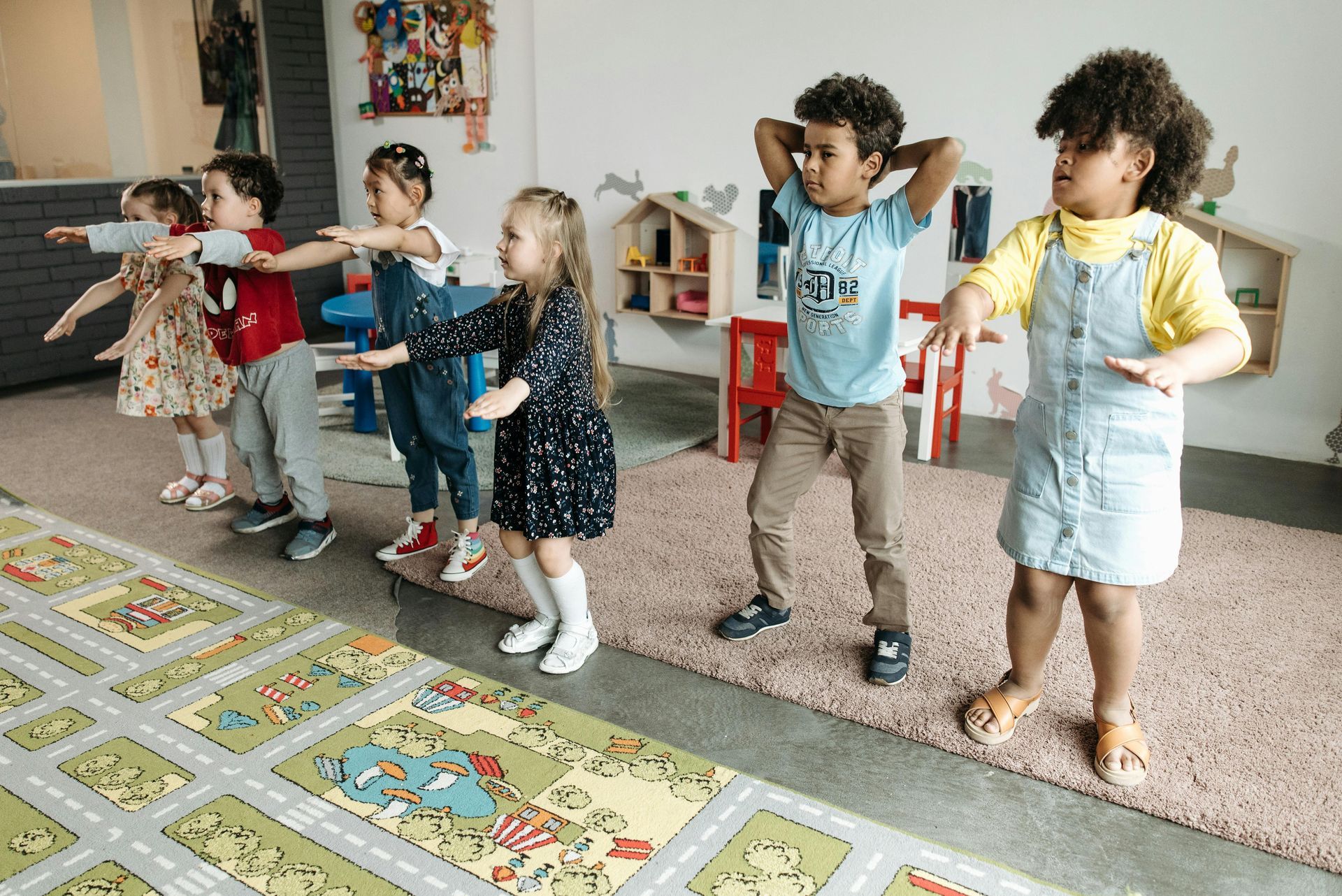 Children dancing in a classroom, colorful clothes, arms outstretched, happy expressions, carpet with city design.