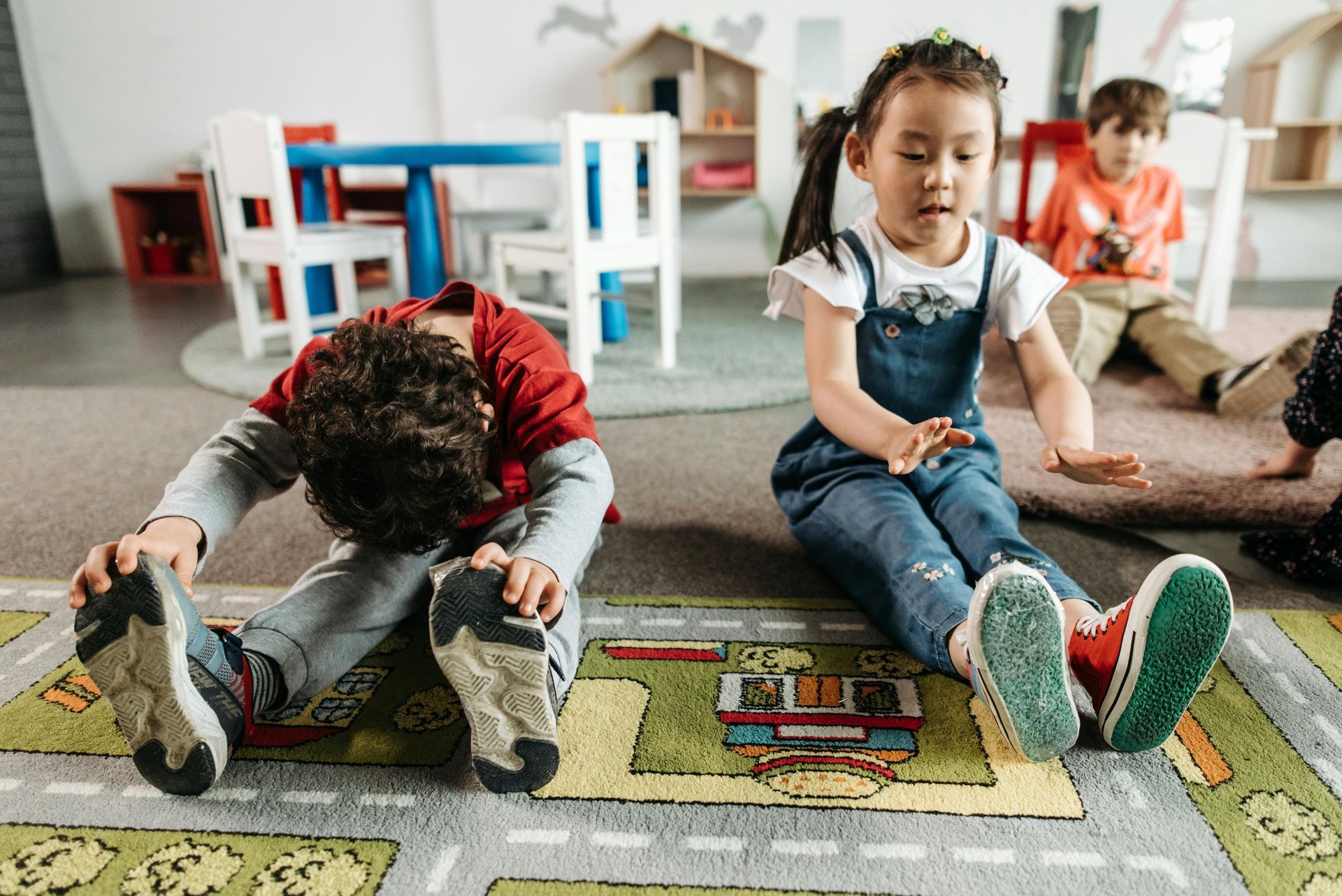 Children stretching on a playroom rug: boy touching toes, girl reaching, others in the background.