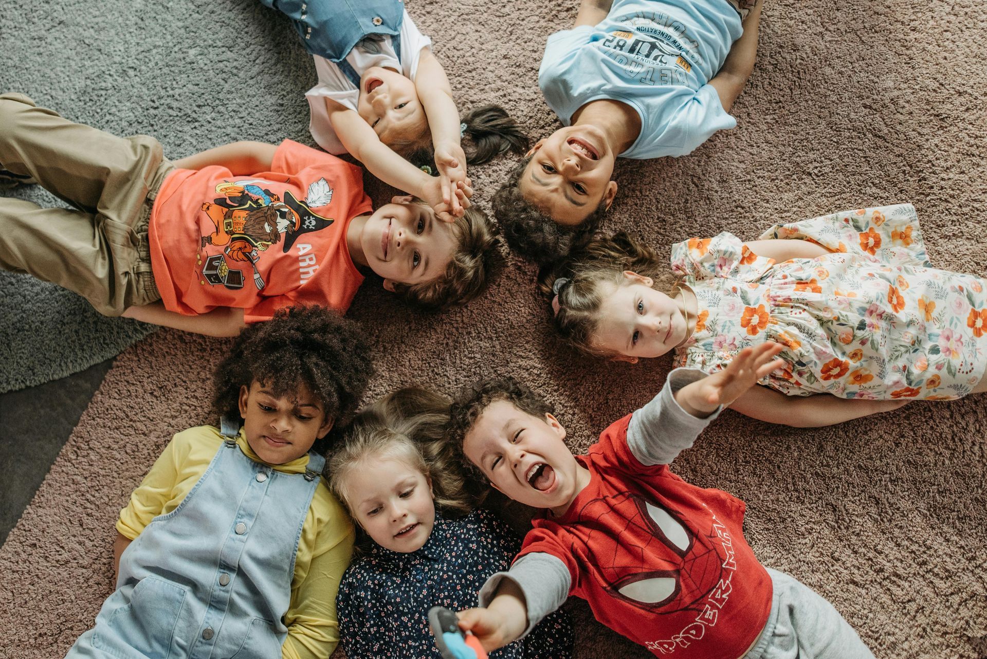 Children lying in a circle on a rug, smiling and looking up at the camera.