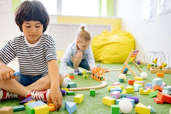 Boy with dark hair and striped shirt playing with colorful blocks on green carpet, girl in background.