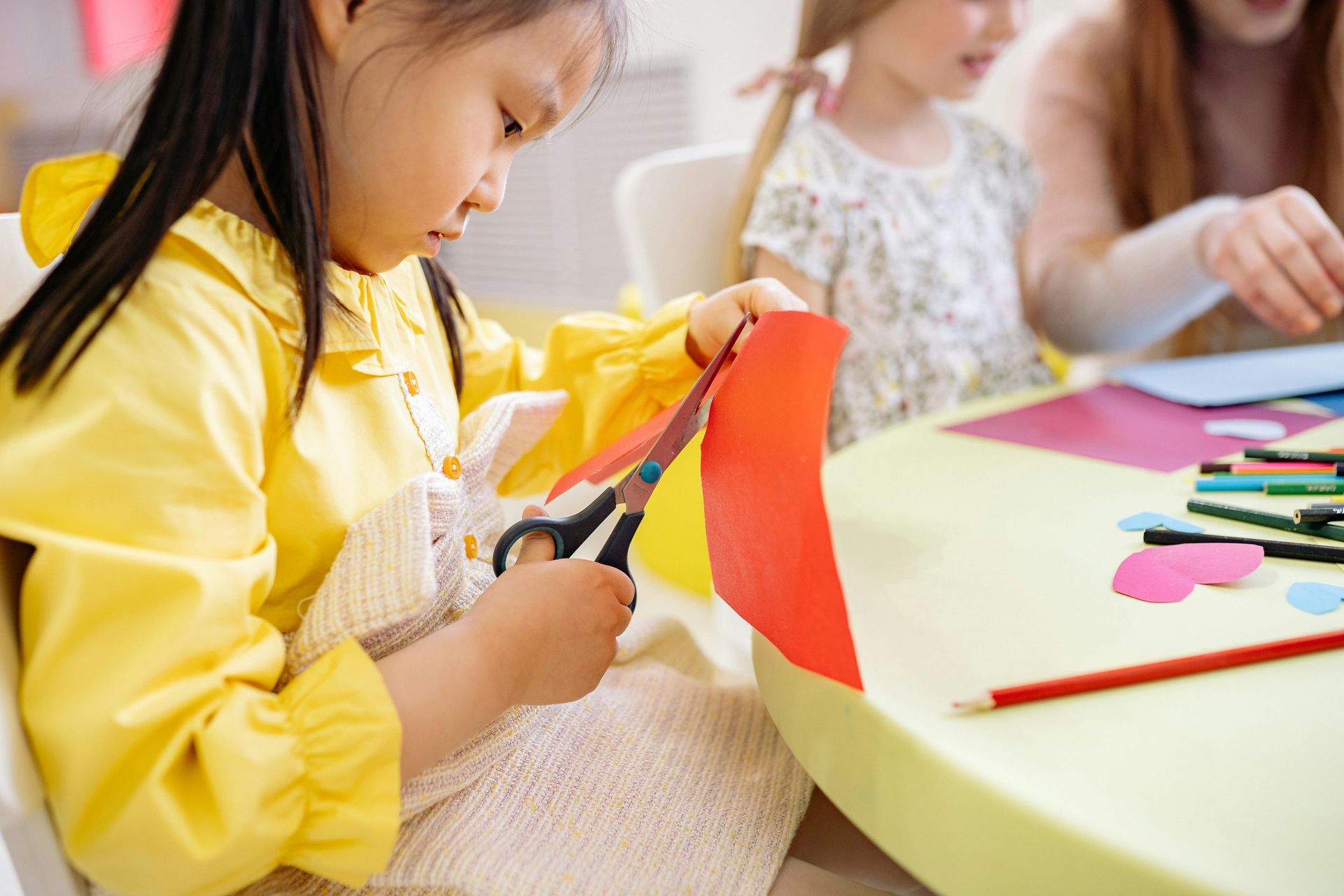 Girl in yellow jacket, cutting red paper with scissors at a table in a classroom setting.