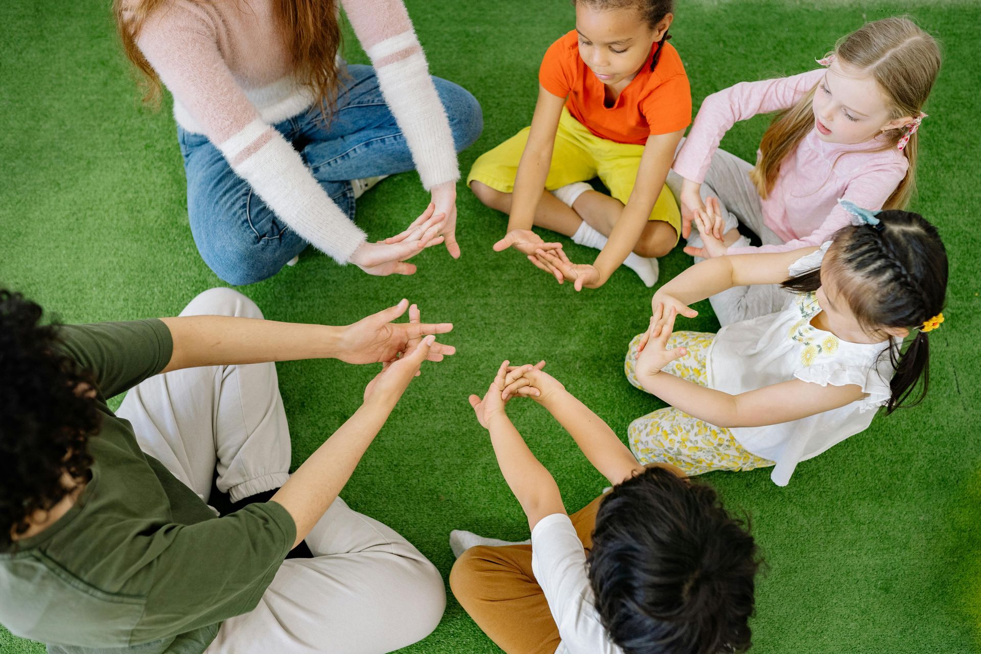A group of children and an adult sit in a circle on green grass, hands touching in the middle.