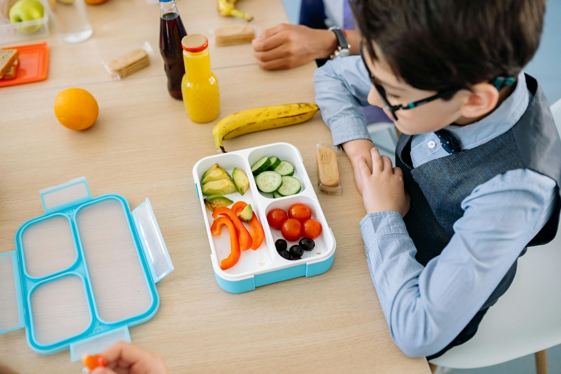 Boy with glasses looking at a lunch box filled with vegetables, on a table with snacks.
