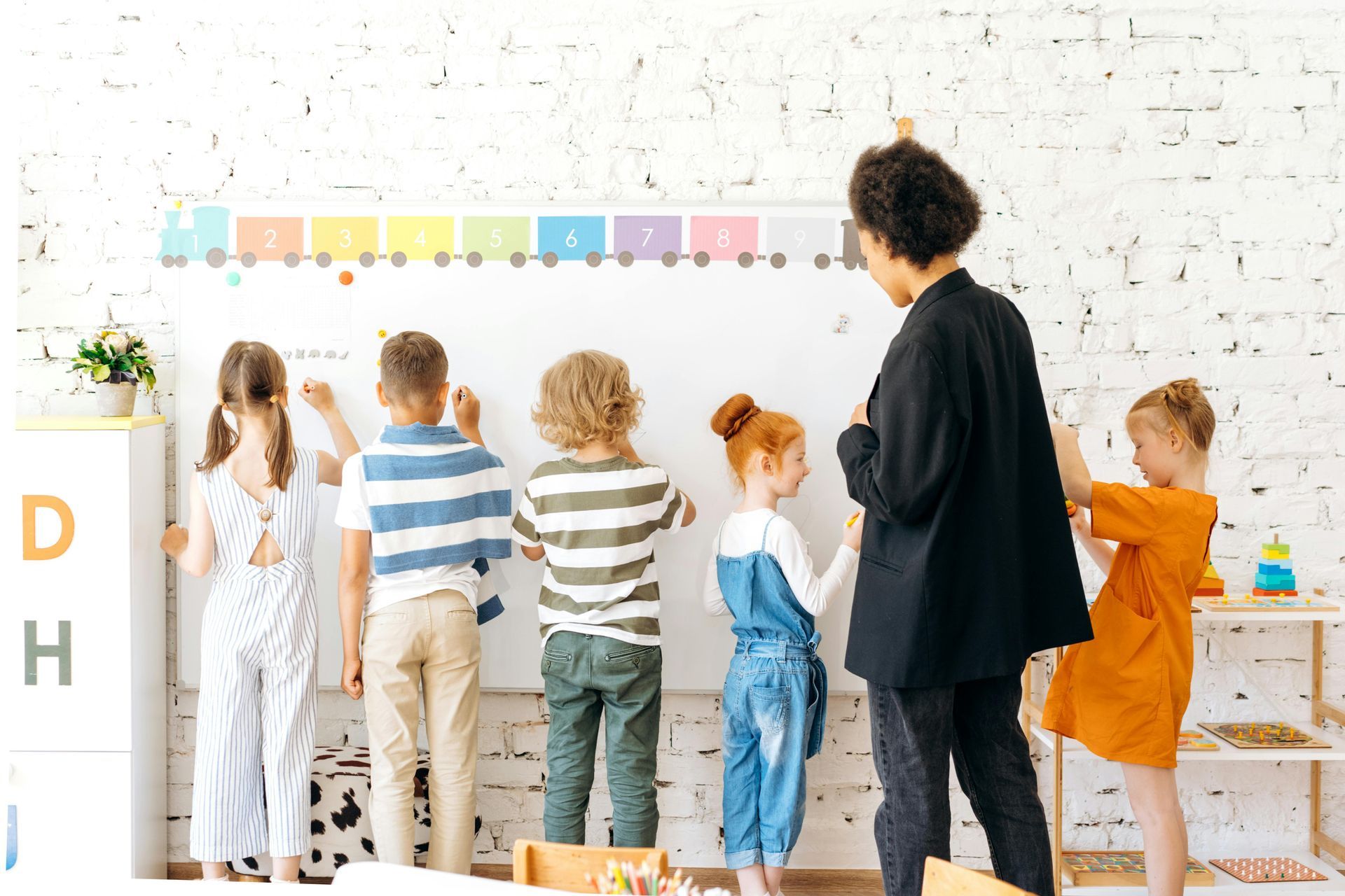 Teacher observes children writing on a whiteboard in a classroom; numbered train banner above.
