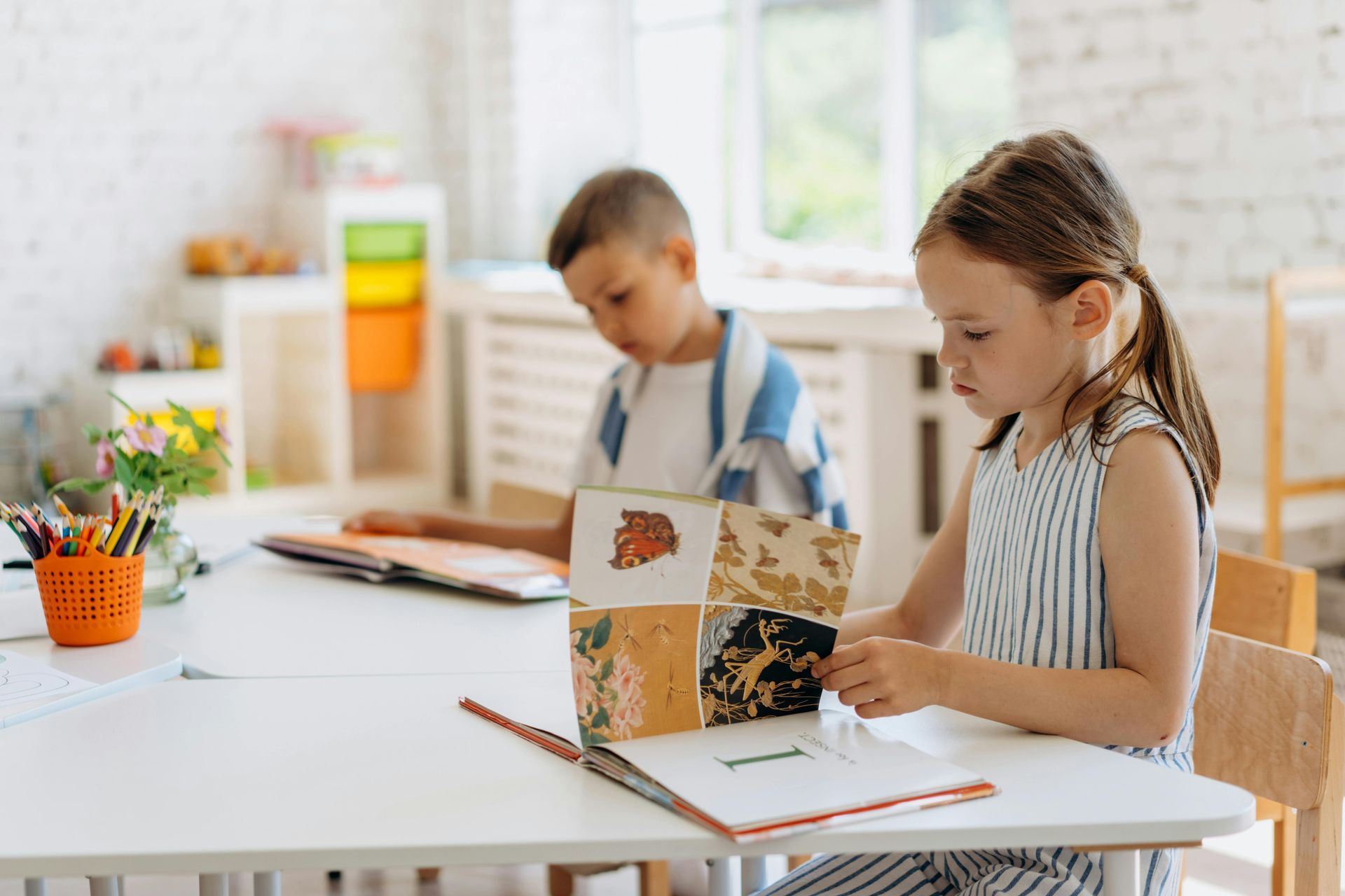 Two children at a white desk; girl in a striped dress looking at a book, boy behind her reading.