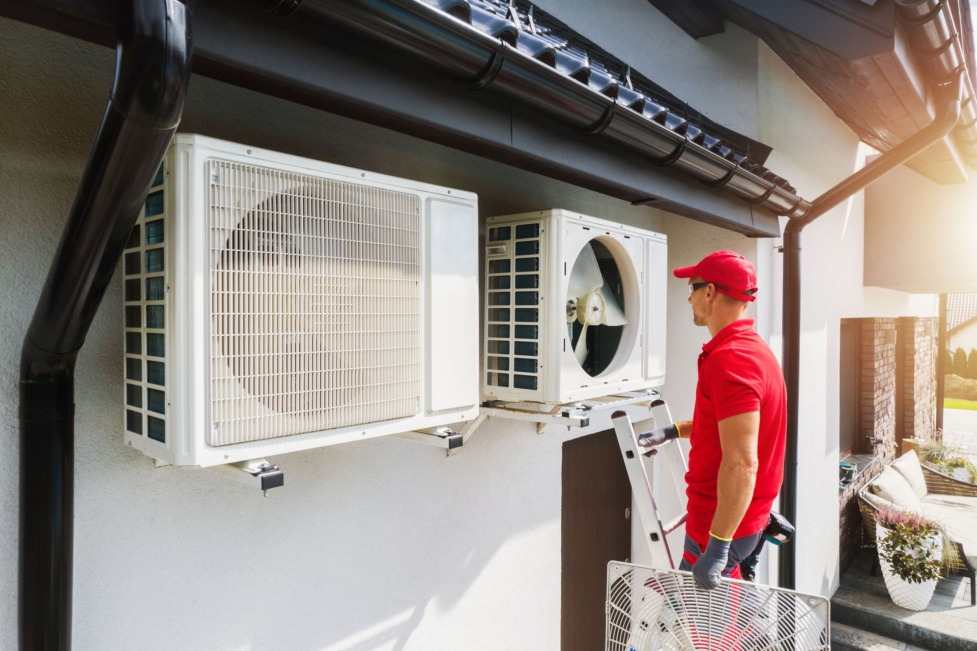 A technician in a red shirt and cap installs an air conditioning unit on a building's exterior.