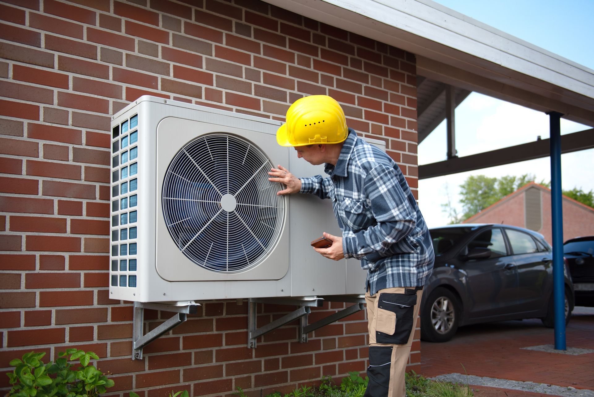 A person in a hard hat inspects an outdoor air conditioning unit mounted on a brick wall next to a carport.