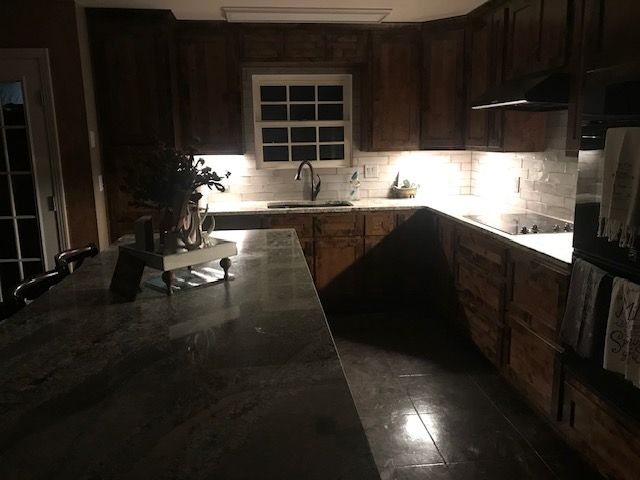 Dark kitchen with brown cabinets, a gray countertop island, and under-cabinet lighting. A window and sink are visible.