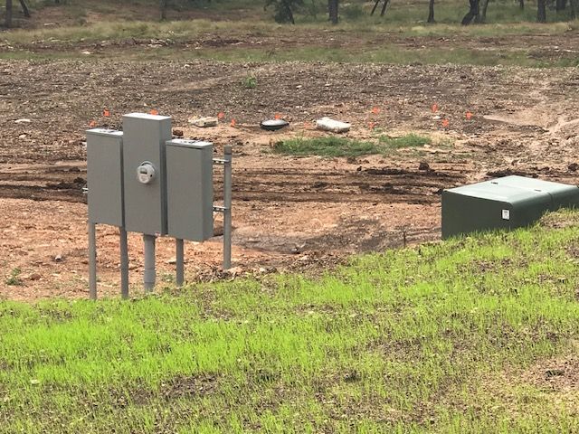 Electrical equipment, including metal boxes and meter, on a grassy slope at a construction site, with dirt and vegetation in the background.