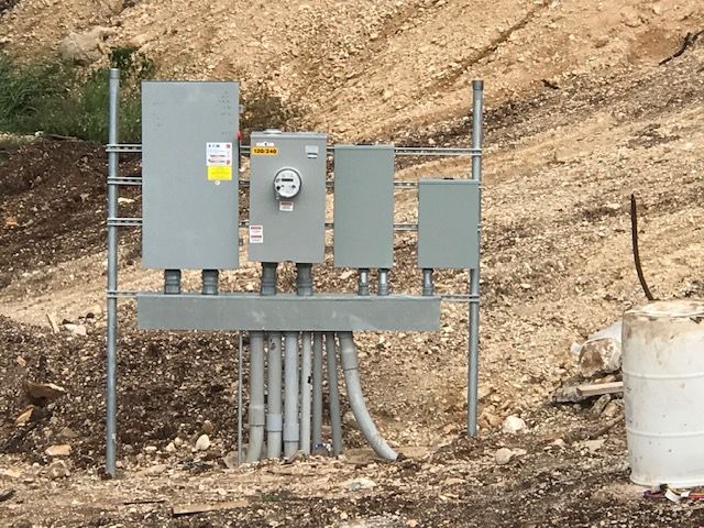 Electrical utility boxes mounted on a metal frame in a dirt setting. Gray boxes with conduit pipes.