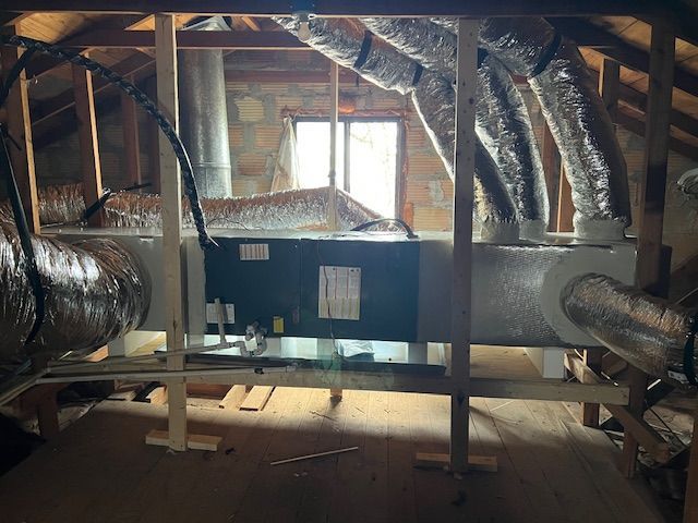 An attic space with an HVAC unit. The unit is black and silver with insulated ductwork. A wooden frame supports the system.