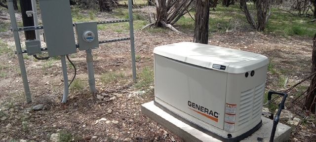 A Generac generator on a concrete pad next to electrical boxes in a wooded area.