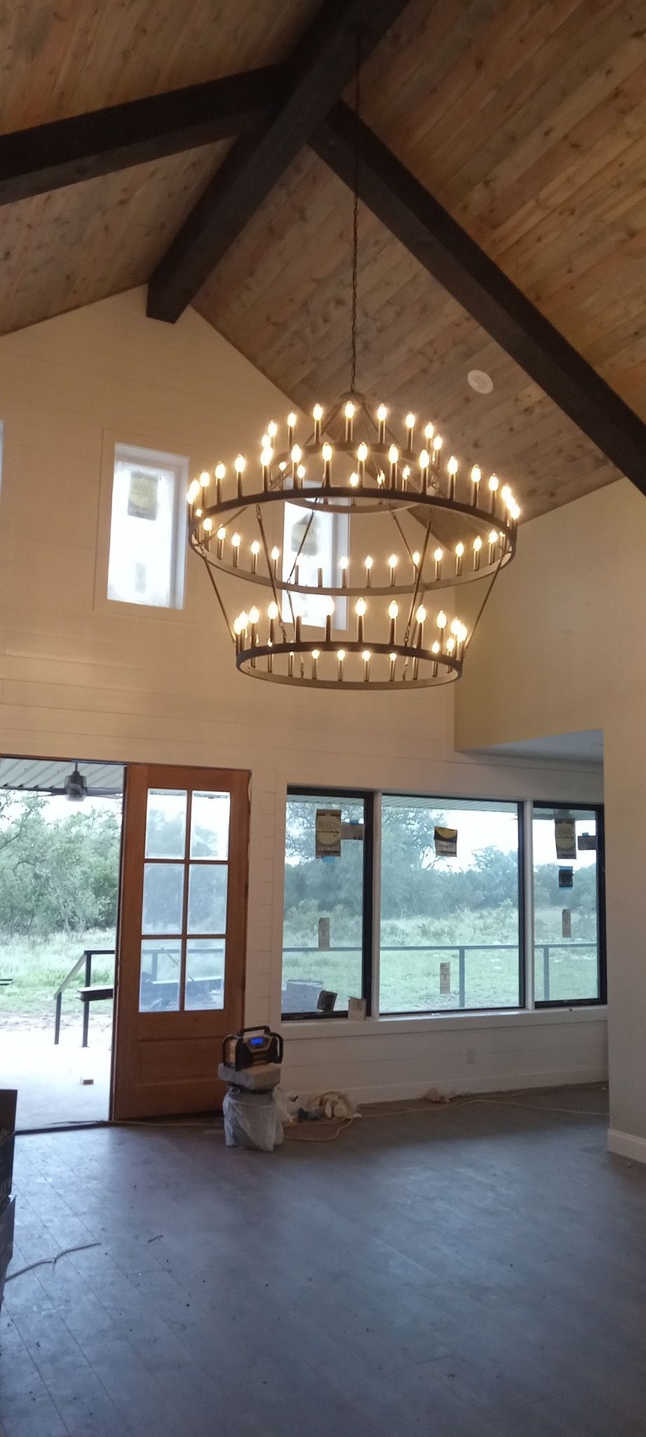 An open-plan interior with a large, tiered chandelier. The room has high ceilings, wood beams, and an outdoor view through windows and a door.
