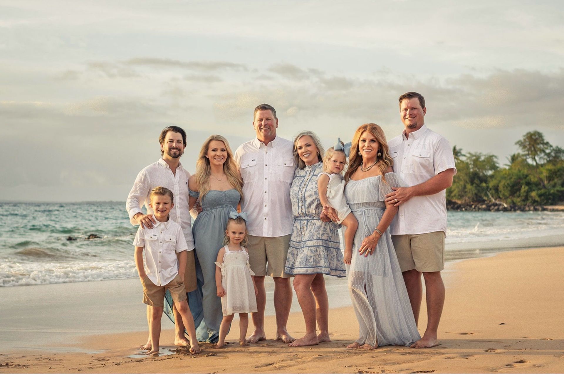 Family of ten poses on a beach at sunset; adults in white or blue and tan, children in light colors, some looking at the camera.