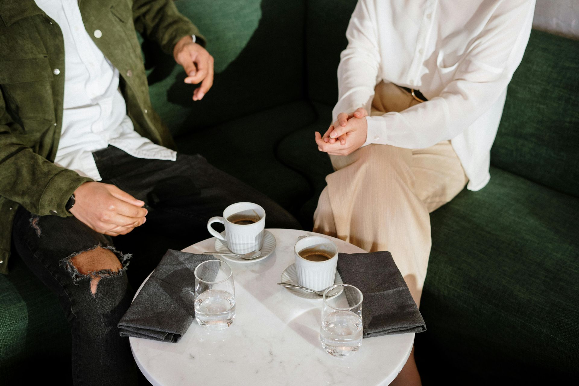 Two people seated, talking, with coffee cups and water glasses on a small table.