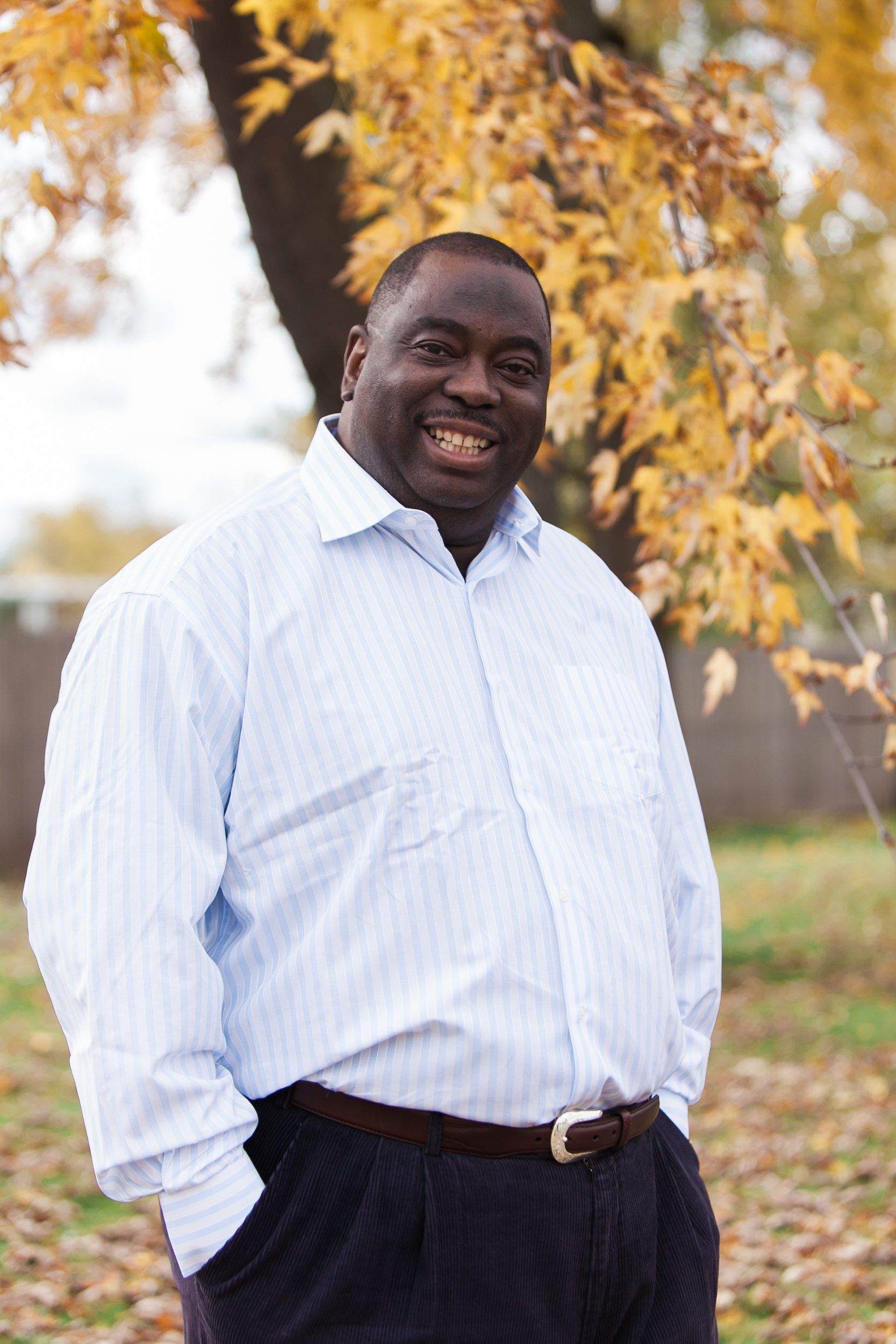 A man in a blue shirt is standing in front of a tree with his hands in his pockets.