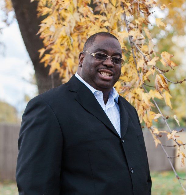 A man in a suit and glasses is smiling in front of a tree