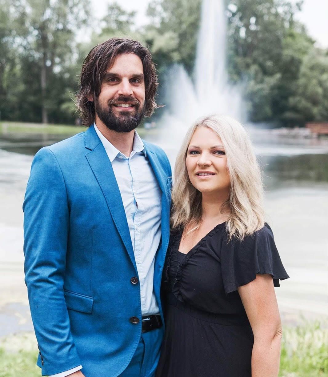 A man in a blue suit and a woman in a black dress are posing for a picture in front of a fountain.