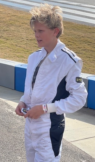 Young person in a white racing suit stands near a track. The suit has black accents.
