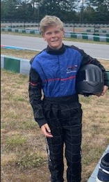 Boy in racing suit and helmet at a racetrack, smiling.