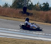 Go-kart racing on a track, driver in white racing suit and helmet.