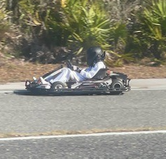 Person in a go-kart wearing a helmet, driving on a road next to foliage.